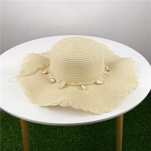 Chapeau de paille beige à bord ondulé avec des accents de coquillages sur table blanche
