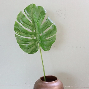 Artificial green Monstera leaf in a brown metallic vase against a minimalist light wall
