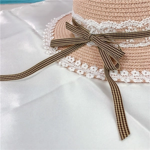 Lace-trimmed straw sun hat with checkered ribbon bow on a white satin fabric backdrop