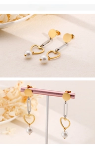 Detail of heart drop earrings featuring gold heart charms and white pearls on a pink stand