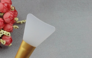 Detail showing a white silicone face mask brush with gold metal handle on a gray surface