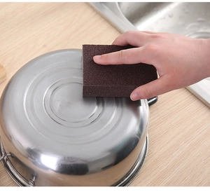 Close-up of brown emery sponge cleaning stubborn black stains from a stainless steel pot