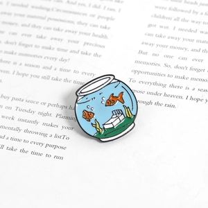 Detail shot of a fish tank enamel pin with orange fish on an open book background