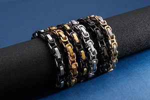 Detail of men's stainless steel box chains in gold, silver, and black on a textured prop