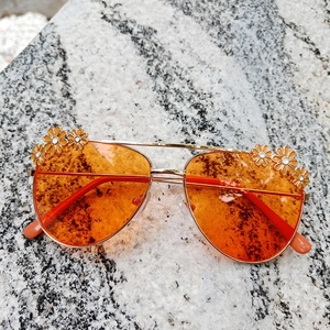 Detail view of gold pilot frame sunglasses with daisy accents and bright orange lenses