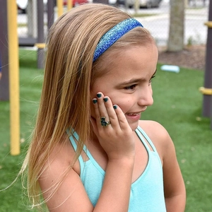 Young girl wearing a blue sequin gradient headband and green toy ring outdoors