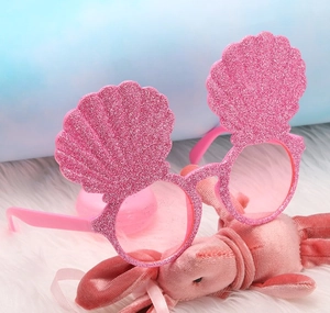 Pink glitter shell shaped sunglasses displayed on white plush background for parties