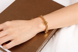 Close-up of a gold heart charm bracelet on a model wrist against a white fabric backdrop