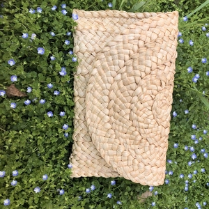 Dettaglio borsa a busta in paglia intrecciata a mano su erba verde con fiori blu