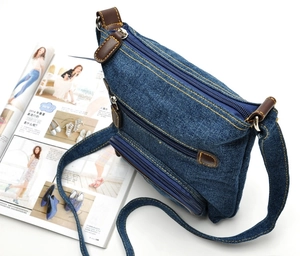 Detail of blue denim crossbody bag with brown leather trim and silver metal zippers