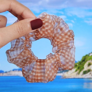 Close-up of brown plaid organza scrunchie held by hand against a blurred beach backdrop