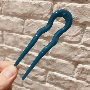 Detail of dark teal wave U-shaped resin hair fork held against a white brick wall background
