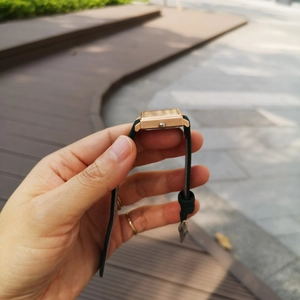 Side view of square gold watch with black leather strap held against outdoor path