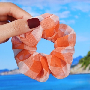Orange and white gingham plaid scrunchie held by hand against a sunny blue sky beach background