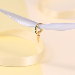Detail of gold huggie earrings with green zircon and sharp spike charm on white ribbon