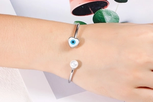 Detail of silver open cuff bracelet with heart evil eye charm and white pearl on wrist