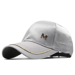 Detail of silver satin baseball cap featuring metal M emblem and gold trim on the brim
