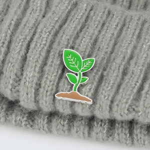 Detail of a green sprout plant enamel pin attached to a grey knitted beanie hat