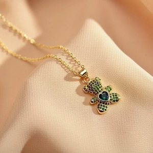 Gold teddy bear necklace with green heart-shaped crystal pendant on beige fabric