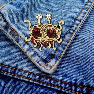 Flying Spaghetti Monster enamel pin accessory on a dark blue denim jacket pocket