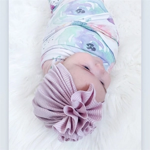 Purple ruffled cotton baby hat with flower accent and floral swaddle on soft plush rug