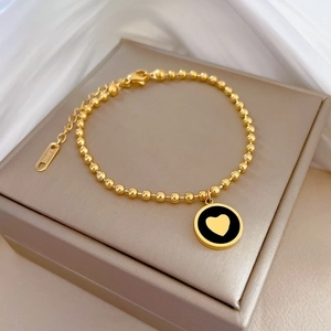 Détail du bracelet chaîne à billes en acier doré avec pendentif cœur sur fond de soie
