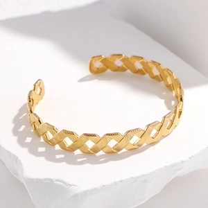Golden geometric hollow cuff bracelet with woven texture on a white stone pedestal
