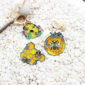 Three detailed bee and honeycomb enamel pins on a natural pebble and shell background