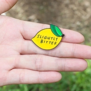 Vibrant yellow lemon enamel pin with green leaf and slightly bitter text on outdoor background