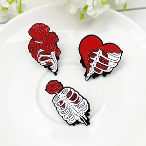 Gothic skull hand and ribcage heart enamel pins with red roses on white ceramic tray
