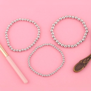 Detalle de conjunto de tres pulseras minimalistas de cuentas plateadas sobre fondo rosa