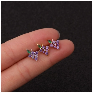 Three tiny gold cartilage studs shaped like purple grapes with a green leaf on a fingertip