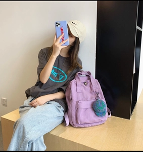 Purple canvas backpack with green hat charm shown in a casual lifestyle setting