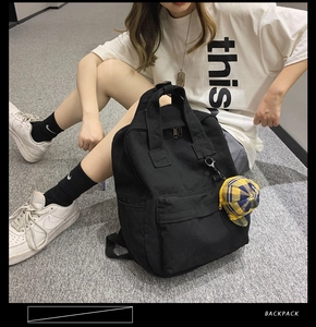 Detail view of a black canvas backpack with a plaid hat charm shown in a lifestyle setting