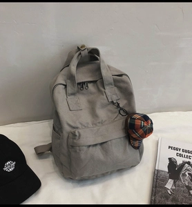 Gray canvas backpack detail showing adjustable straps, top handle, and checkered hat pendant