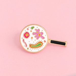 Cartoon enamel pin featuring a colorful microbe cell badge under a magnifying glass design