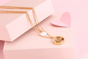 Detail view of a double-layered gold necklace with a Mum pendant on a soft pink background