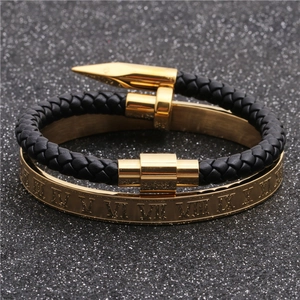 Close-up of gold Roman numeral bangle and black braided nail bracelet set on grey surface