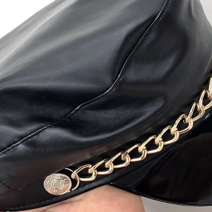 Detail of black faux leather newsboy cap featuring a bold gold chain and silver star button