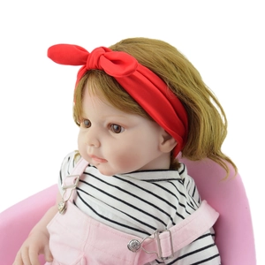 Realistic baby doll with brown hair, red bow headband and pink overall shorts