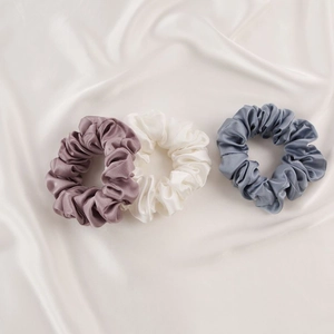 Three premium mulberry silk scrunchies in purple, cream, and blue on a soft satin backdrop