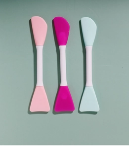 Three dual-ended silicone face mask brushes in pink, rose, and blue on a green base