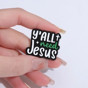 Black metal enamel pin with y'ALL need Jesus text shown in a hand-held close-up shot