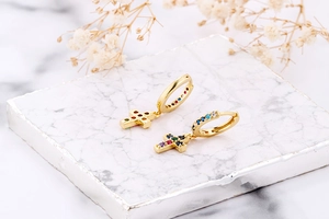 Detail of gold huggie earrings with rainbow zircon cross charms on marble surface
