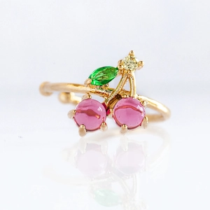 Gold-plated cherry ring featuring pink fruit and green leaves on a white background