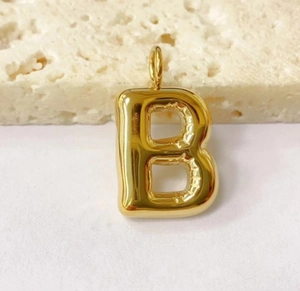 Close-up of a golden bubble letter B pendant resting on a textured beige stone background