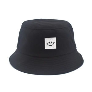 Detail of a black cotton bucket hat featuring a white square smiley face patch on the front