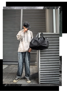 Male model holding black fabric duffel bag against a metallic wall in street style shot
