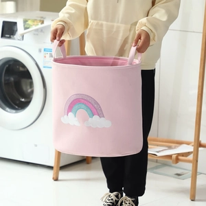 Pink round laundry basket with rainbow embroidery and handles held in a laundry room