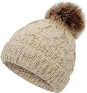 Close-up of beige knit winter hat featuring chunky cable patterns and a large faux fur pompom